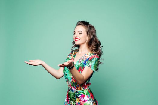 Studio portrait of beautiful smiling brunette with long wavy hair in floral summer dress showing two things on her palm hands over green background.