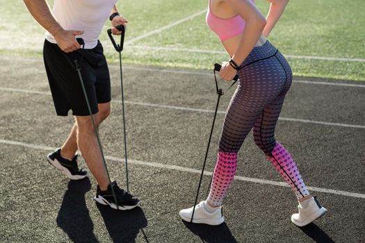 Close-up of a young man and woman training with an expander at the stadium in the summer, pumping the muscles of the back and arms