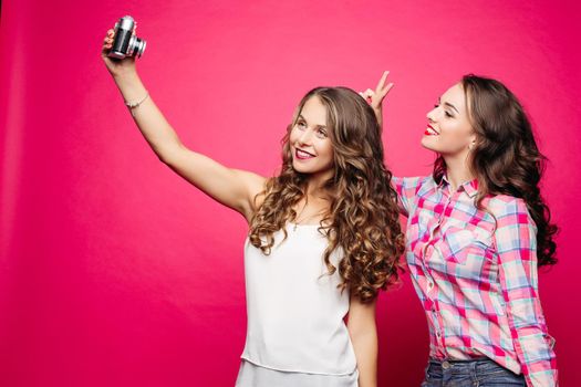 Portrait of happy attractive girlfriends with long wavy hairstyles taking self-portrait via old film camera against red background. Brunette in plaid shirt making bunny ears to her friend.