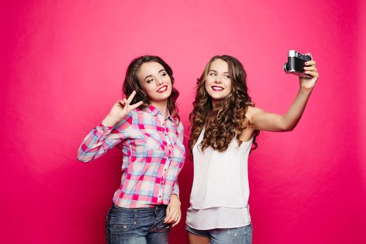 Portrait of happy attractive girlfriends with long wavy hairstyles taking self-portrait via old film camera against red background. Brunette in plaid shirt making bunny ears to her friend.