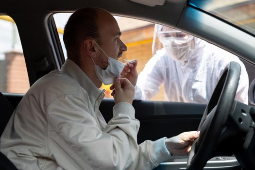 A man wearing a protective suit, gloves, surgical mask, and face mask is testing the coronavirus co id-19 on another man sitting in a car.