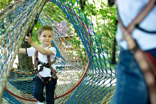 Portrait of a little boy in climbing gear in a rope park, holding a rope with a carbine.