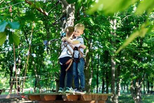 Two cute boys in an adventure park are doing rock climbing or passing obstacles on a rope road, hugging