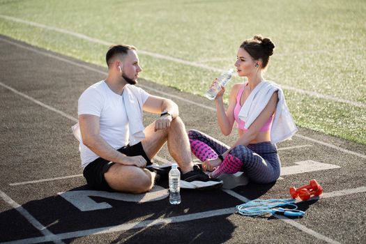 Man and a woman sit on the treadmills after a run at the stadium in the summer, they listen to music with headphones