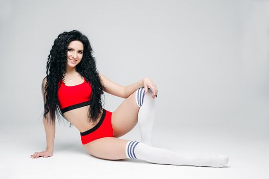 Full lenght portait of sexy dark haired girl wearing in red sports wear and white leggings lying on floor. Woman posing at white studio background, smiling at camera. Concept of sport, fitness, dance.