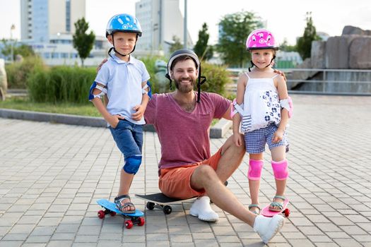 Father with his children, son and daughter, skate in the park, smile and look at the camera.