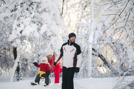 Winter walk: parents sledding with their son. A father pulls a sled with a young son on a snowy day, and a mother pushes.