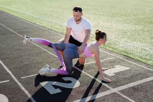 Coach plays sports with a young girl at the stadium. Leg exercise with elastic band