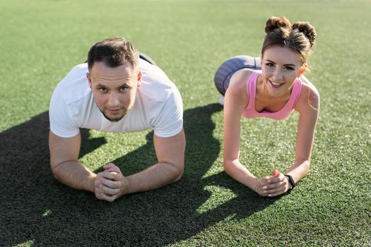 Close-up of a man and a woman doing a plank exercise on the grass at a sports stadium at sunset