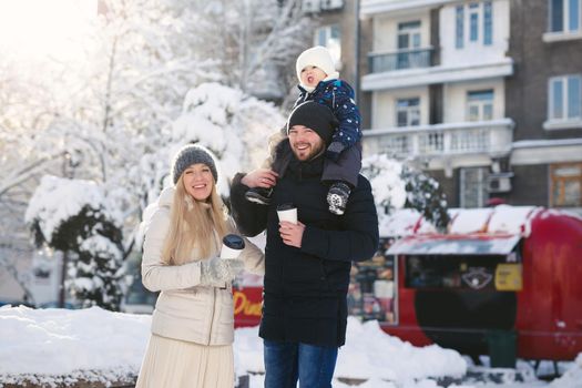 Portrait of a happy young family walking together in a winter city park, sunlight. The son is sitting on Dad's neck.