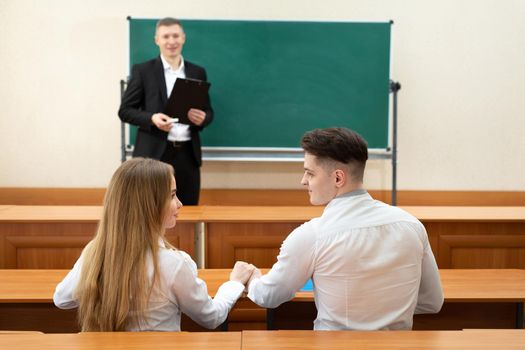 Couple of students in love are sitting at a desk, holding hands and looking at each other against the background of the teacher.