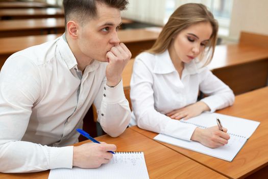 Young couple of students, a guy and a girl, sit at a desk at a lecture and hold hands.