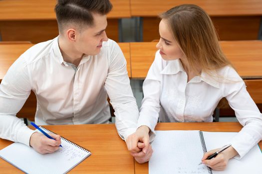 Young couple of students, a guy and a girl, sit at a desk at a lecture and hold hands.