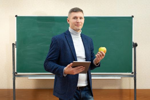Portrait of a smiling teacher with a tablet computer and a green apple in the classroom