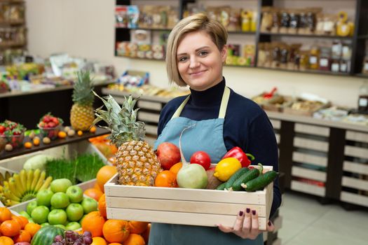 Smiling woman salesman holds a wooden box with vegetables and fruits in the store.
