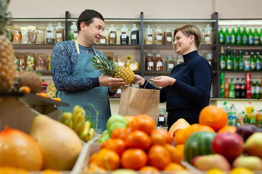 Male salesman passes a craft bag of groceries to a female customer in a store with vegetables and fruits.