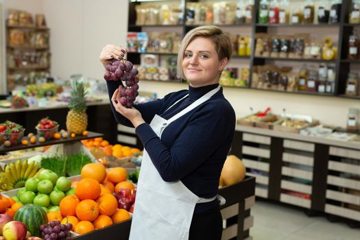 Female saleswoman helps a customer buy fruits and vegetables at a grocery store, holds them in her hands and shows them grapes.