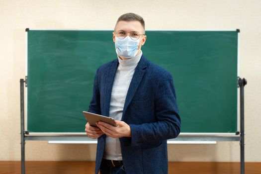 A male teacher in a medical mask stands at the blackboard in the classroom with a tablet in his hands. The concept of a pandemic. School during the coronavirus.