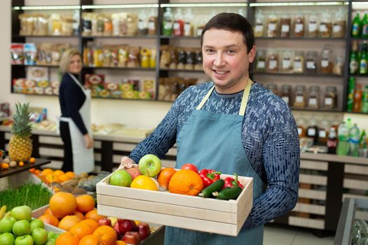 Smiling male salesman holds a wooden box with vegetables and fruits in the store.