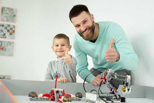 Happy handsome man smiles at the camera with his cheerful little son while playing with lego robots and shows the thumbs up.