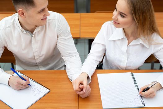 Young couple of students, a guy and a girl, sit at a desk at a lecture and hold hands.