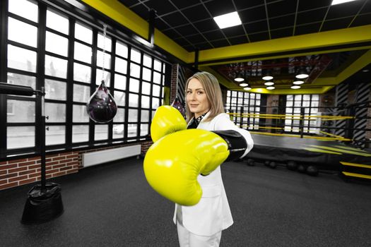 Determined, stylish businesswoman in yellow boxing gloves throws a punch at the camera against the backdrop of a boxing ring.
