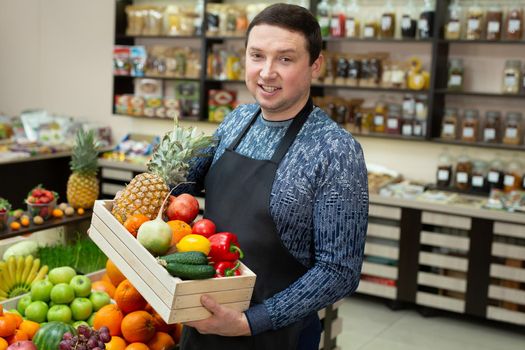 Smiling male salesman holds a wooden box with vegetables and fruits in the store.