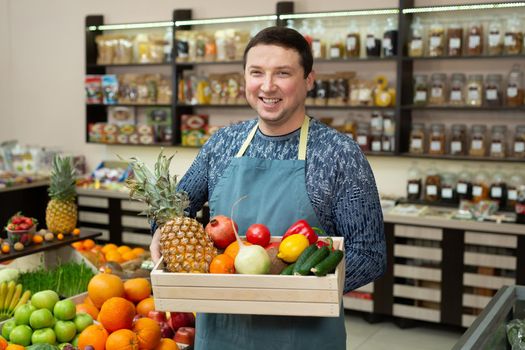 Smiling male salesman holds a wooden box with vegetables and fruits in the store.