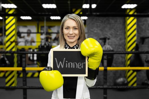 Young businesswoman in a suit in a boxing ring wearing gloves holds a sign with the inscription winner.