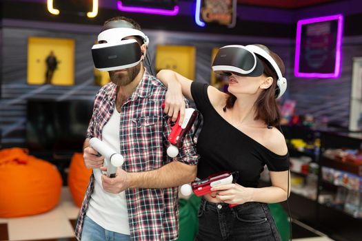 Friends, a man and a woman use a virtual reality headset with glasses and hand motion controllers and weapons in the play area.