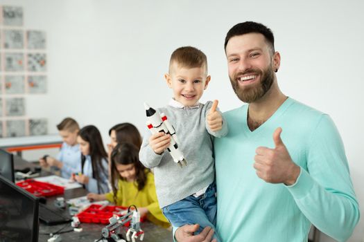 Portrait of a young father with his son in his arms shows the thumbs up in a robotics class.
