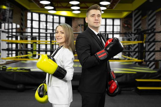 Man and woman in a suit and boxing gloves stand in front of the ring with their backs to each other.