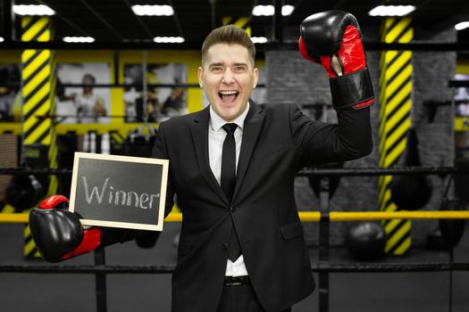 Young businessman in a suit in a boxing ring wearing gloves holds a sign with the inscription winner.