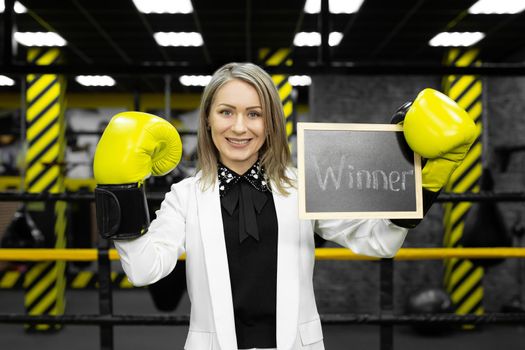 Young businesswoman in a suit in a boxing ring wearing gloves holds a sign with the inscription winner.