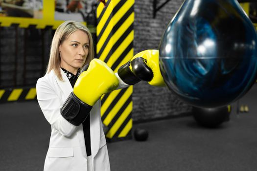 Young female businesswoman in a white suit hits a punching bag in the gym. The concept of office employee anger and relaxation.