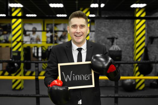 Young businessman in a suit in a boxing ring wearing gloves holds a sign with the inscription winner.