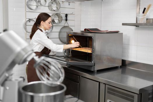 Female pastry chef puts the sponge dough in the oven and checks the dough with a wooden stick, a skewer.