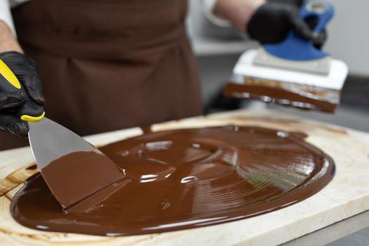 Male chocolatier uses a spatula to stir the tempered liquid chocolate on a granite table