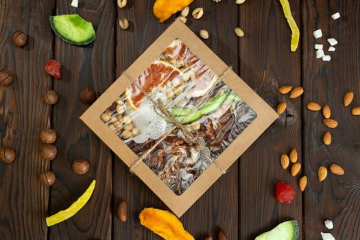 Nuts, candied fruits and dried fruits of different varieties in a paper box on a structural wooden background top view.
