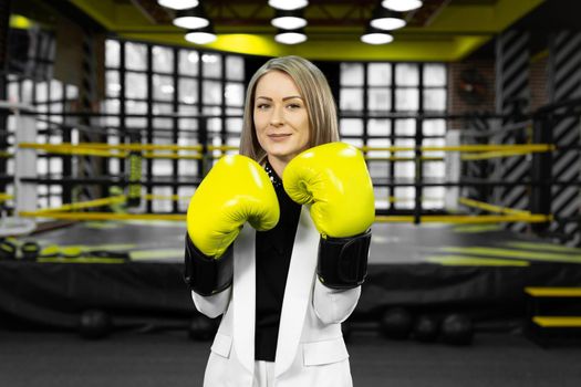 Determined, stylish businesswoman in yellow boxing gloves throws a punch at the camera against the backdrop of a boxing ring.