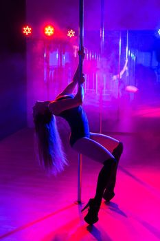 Pole dance. Slender sexy woman dancing in the interior of a nightclub with a colored background and smoke.