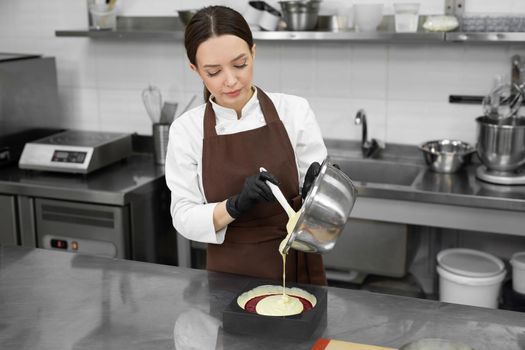 Young female pastry chef pours liquid mousse into a mold. Preparation of cake, dessert in a professional kitchen.