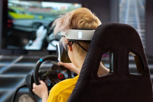 Teenage boy wearing virtual reality glasses, who holds on to the steering wheel and plays a computer game on the console