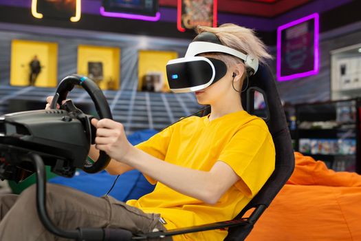 Teenage boy wearing virtual reality glasses, who holds on to the steering wheel and plays a computer game on the console