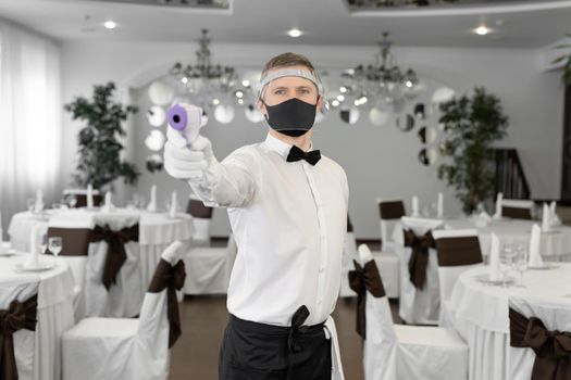 Waitress man wearing a face mask and holding an infrared thermometer on her forehead to check the body temperature for symptoms of the virus