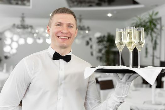 Smiling male waiter with glasses of white wine on a tray in a restaurant.