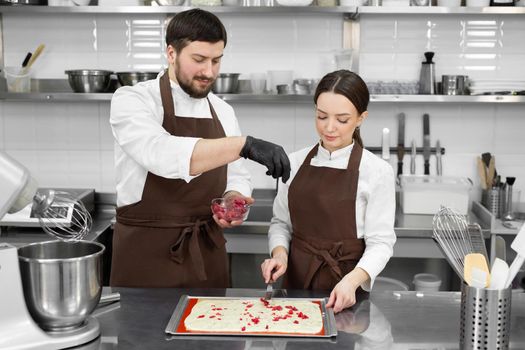 Couple of confectioners, a man and a woman, cook in a professional kitchen and add berries to the sponge cake.