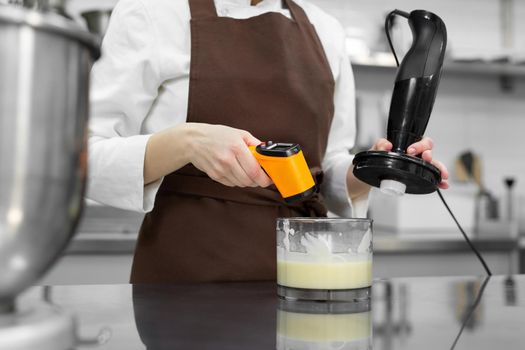 Female pastry chef prepares a mirror icing for a cake, whips it in a blender and measures the temperature.