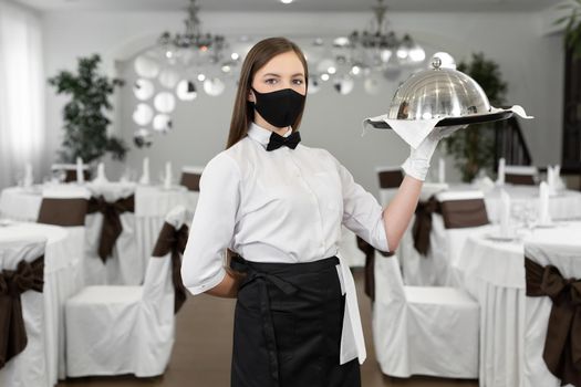 Female waiter in a protective medical mask holds a closed tray with a hot dish.