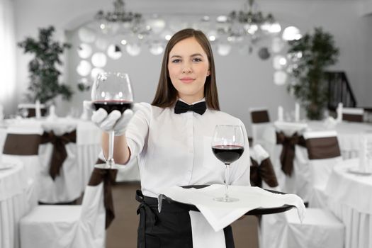 Female waiter in white gloves holds a glass and a bottle of red wine.